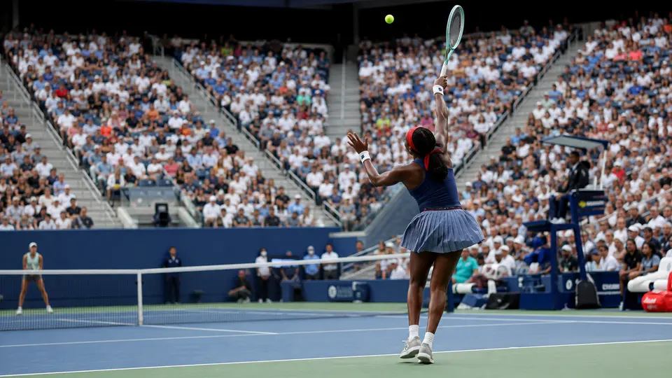 Coco Gauff serving under the closed roof of Arthur Ashe Stadium during her US Open first-round match against Zeynep Sonmez in rainy conditions.